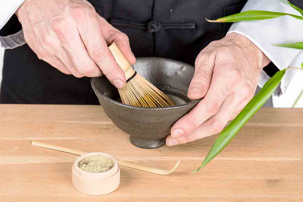 man whisking matcha tea in a mall brown bowl using a bamboo whisk with a small jar or tea and a wooden spoon on a wooden table