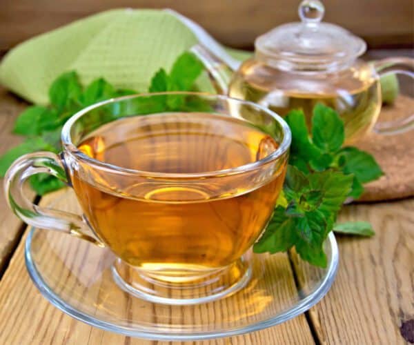 glass tea cup and saucer with a glass teapot containing peppermint tea on a wooden table surrounded by peppermint leaves and a green napkin