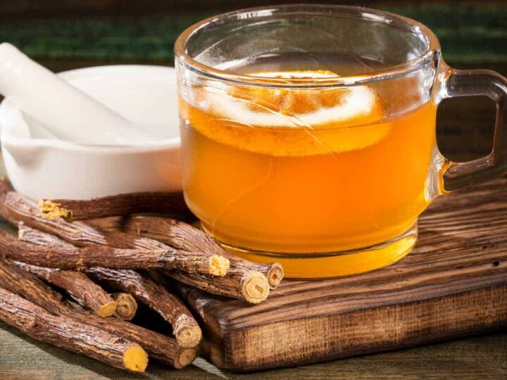 Licorice root tea in a glass cup with an orange slice sitting on a wooden board surrounded with licorice roots and a white mortar and pestle.