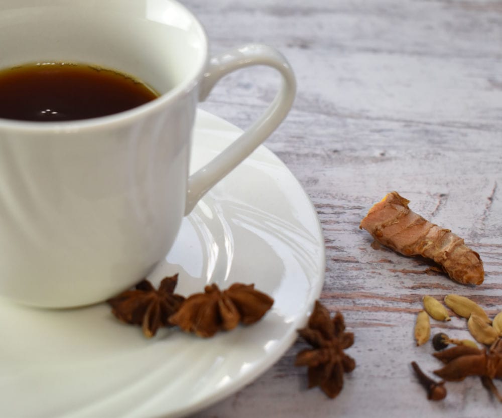 Golden chai in a white cup on a wooden board with spices scattered around.
