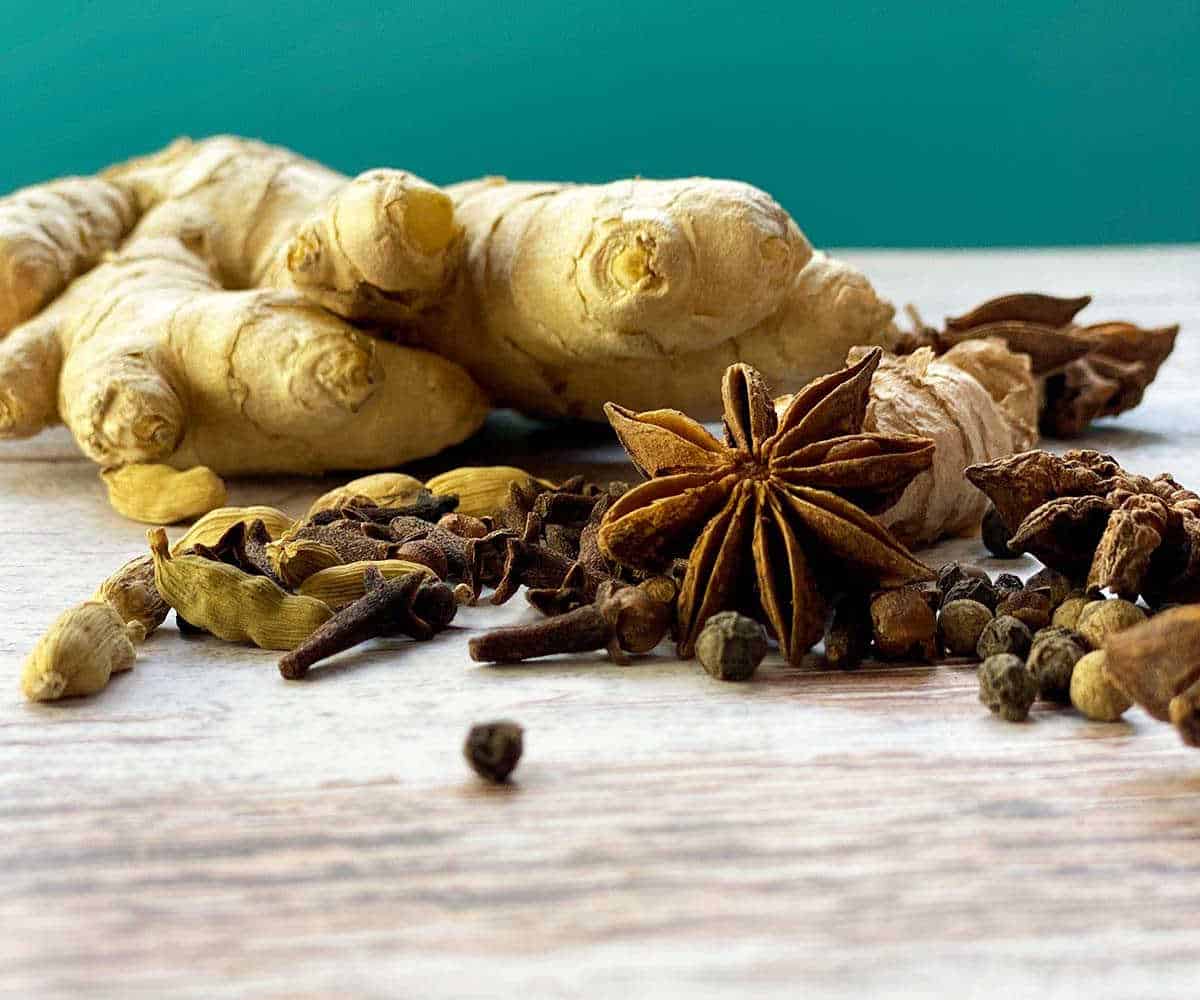 Golden chai spices scattered on a wooden table. Spices include turmeric root, ginger, cloves, peppercorns, cardamom pods, and star anise.
