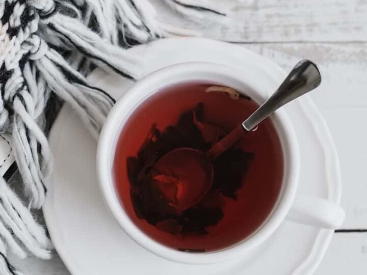 A white cup and saucer containing hibiscus rose hip tea and a spoon on a white wooden background