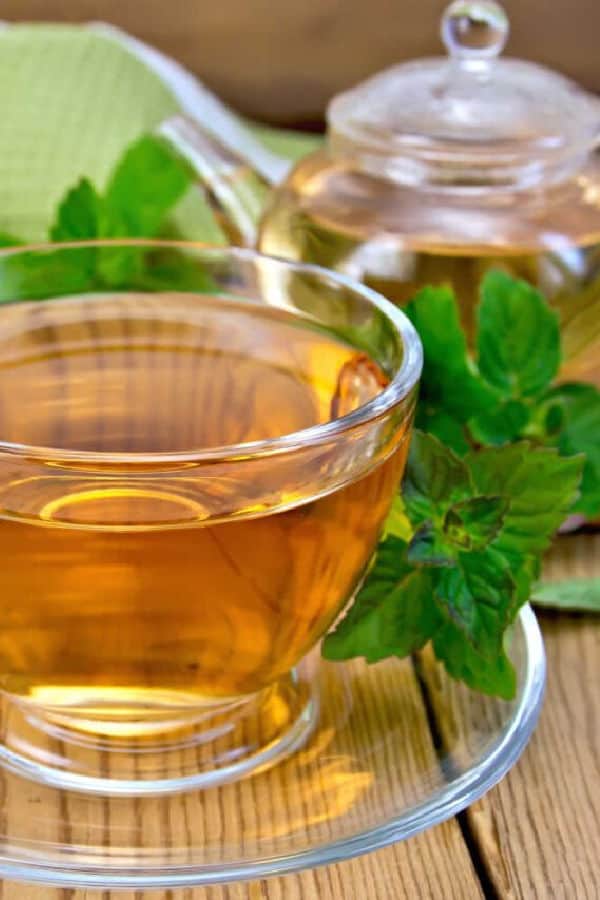 A clear glass tea cup and saucer containing peppermint tea with a peppermint leaf garnish on a wooden table.