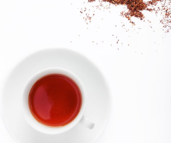A white teacup and saucer containing vanilla rooibos tea with rooibos tea leaves on a white background.