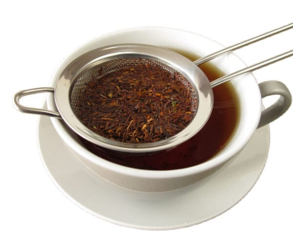 A white teacup and saucer containing vanilla rooibos tea with rooibos tea leaves in a tea strainer on a white background.