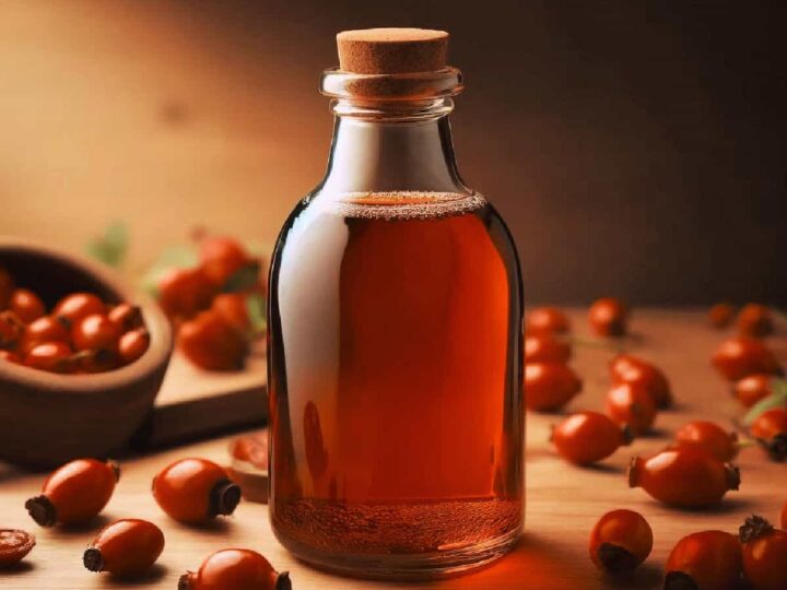 A glass jar containing rosehip syrup surrounded by rosehips.