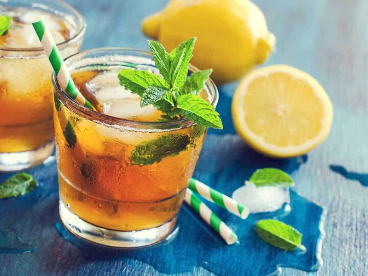 Two glasses of peppermint iced tea on a blue background with lemons, peppermint leaves and two green and white straws.