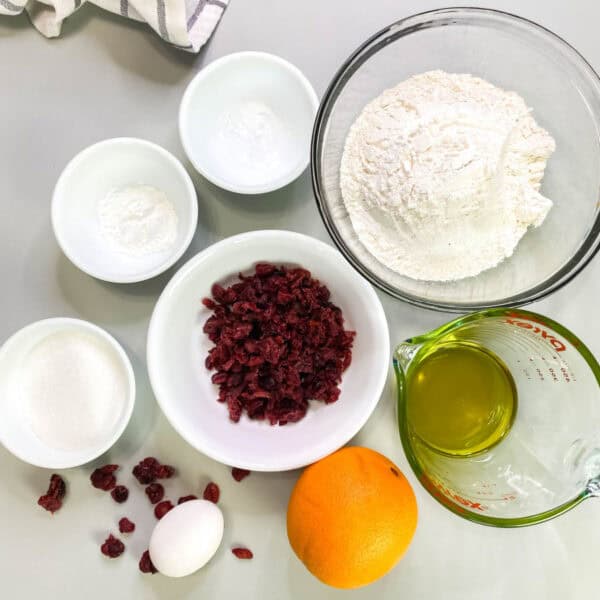 Muffin ingredients, flower, sugar, baking powder, dried cranberries, oil, an orange, and an egg on a white background.