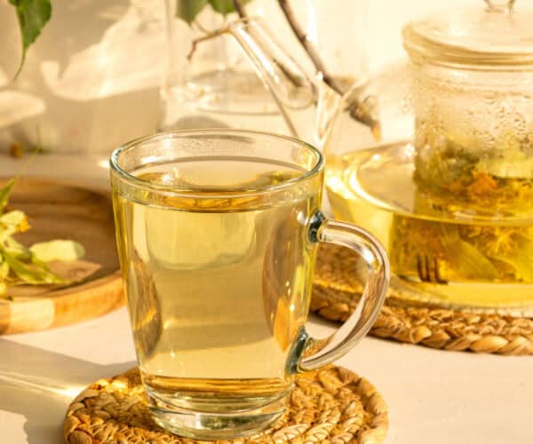 Feverfew tea in a glass tea cup on a rattan coaster with a glass teapot in the background.