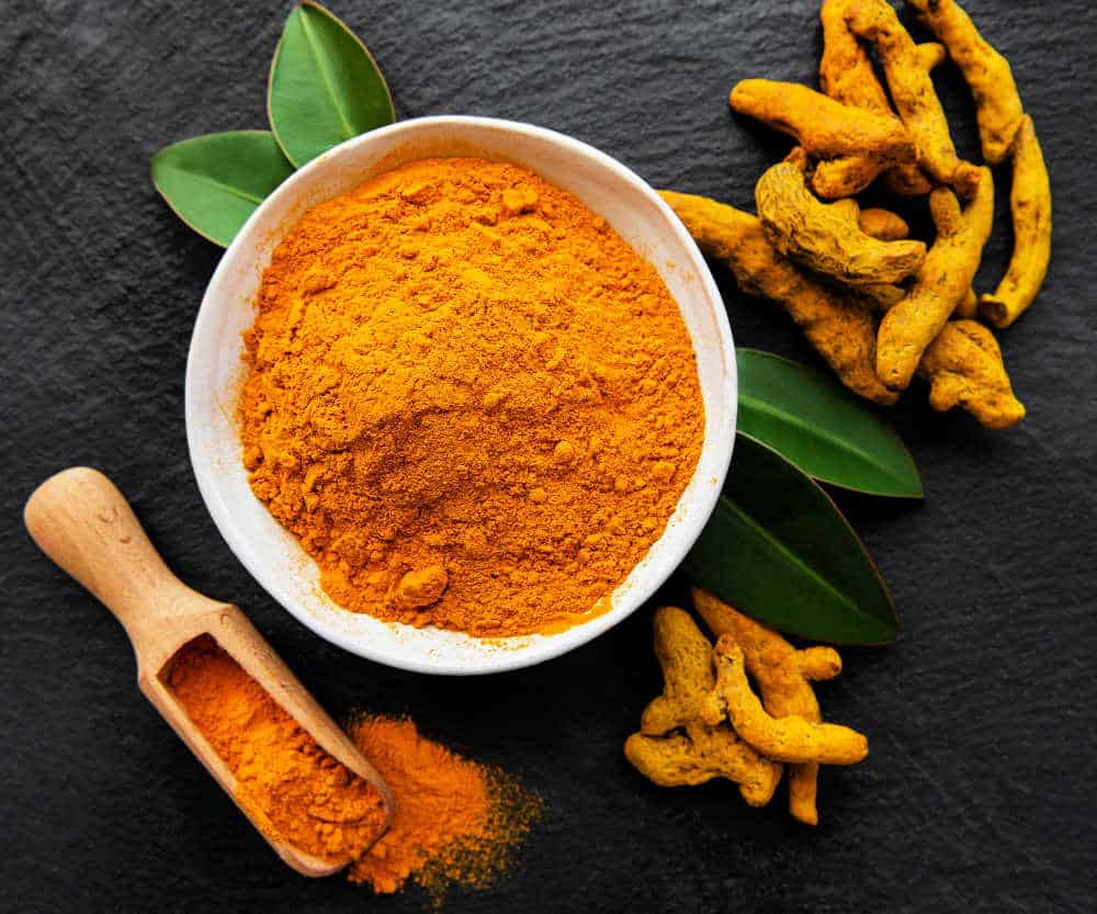 A white bowl containing turmeric powder surrounded by turmeric roots and a wooden scoop filled with turmeric powder.