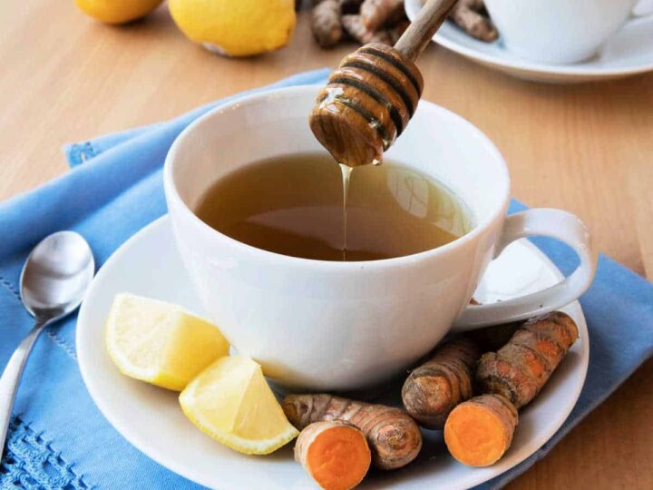 Turmeric tea in a white cup an saucer on a blue cloth surrounded by lemon and turmeric root.