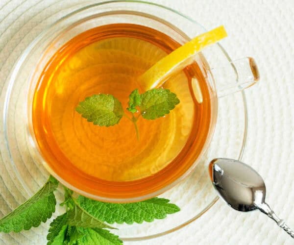 Lemon balm tea in a glass cup and saucer with green leaves and a lemon wedge.