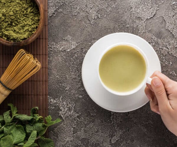 A hand holding a teacup with matcha mint tea on a stone background. There is a wooden bowl of matcha powder, a bamboo whisk and mint leaves on a bamboo mat near by.