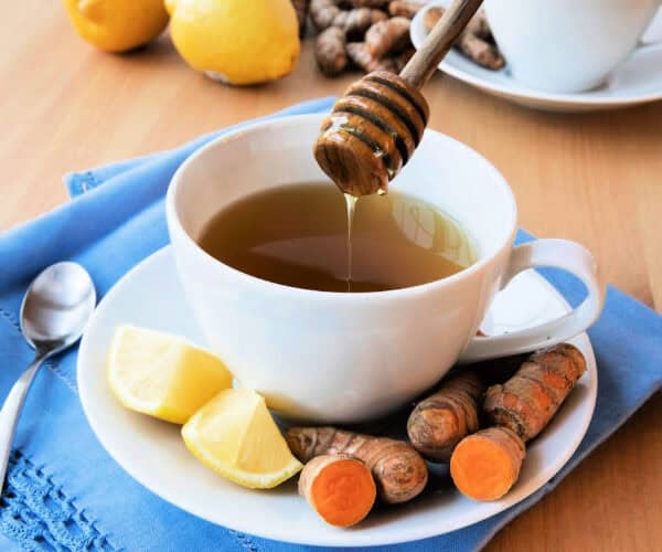 Turmeric and Ginger tea in a white cup and saucer on a blue napkin with turmeric root and lemon and honey dripping from a wooden server.