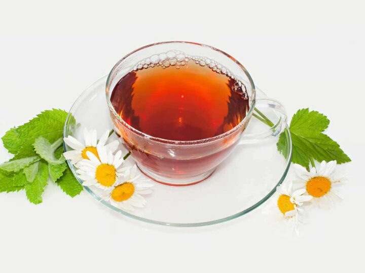 A glass teacup and saucer with tea and chamomile flowers and mint leaves.