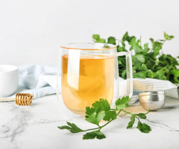 Parsley tea in a glass cup surrounded by a tea ball, a dish of honey, and fresh parsley.