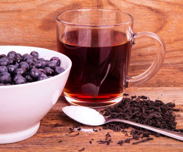 Blueberries in a white bowl. A glass cup with tea, a spoon with sugar, and loose leaf tea on a wooden background.