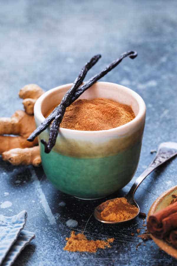 Chai latte in a ceramic bowl with vanilla bean pods and fresh ginger on a dark textured surface.