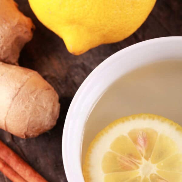 Lemon slice in a white dish with ginger, cinnamon sticks, and a lemon on the side.