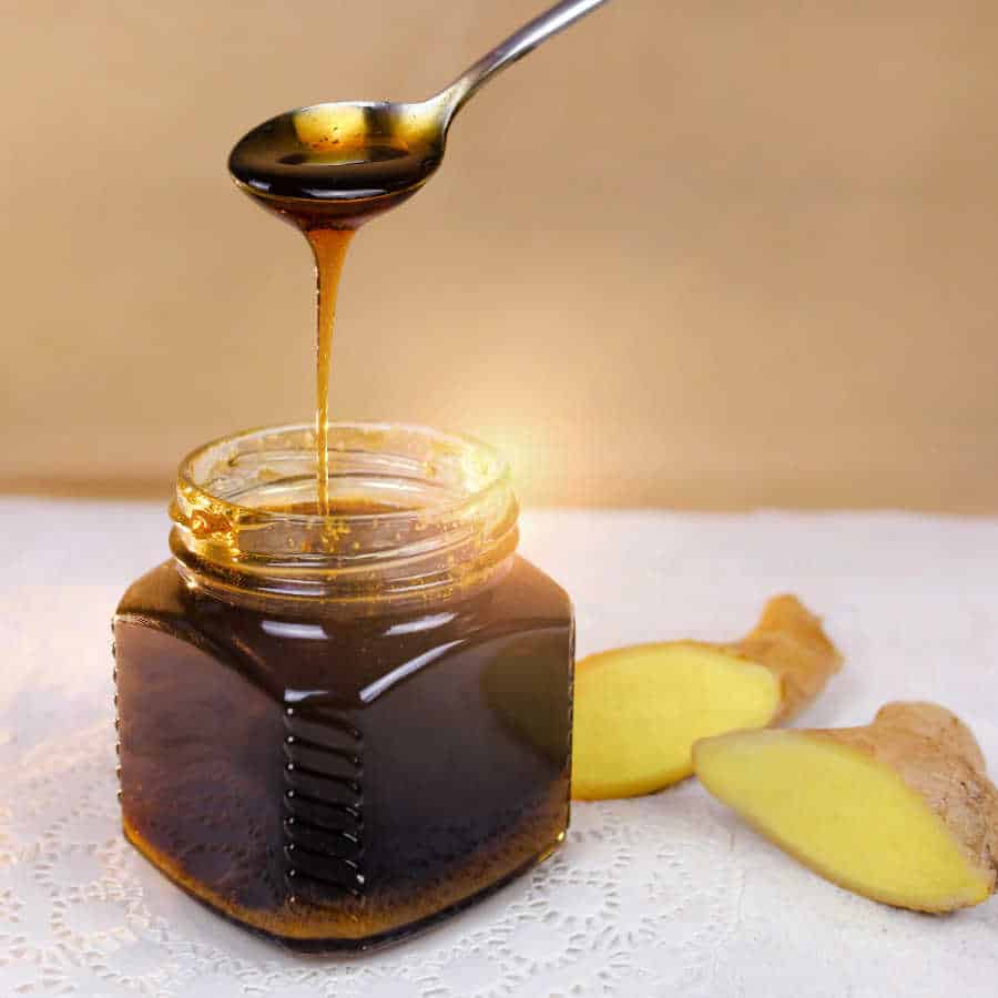 A small glass jar of gingerbread syrup with a spoon dripping syrup into the jar and ginger slices near by.